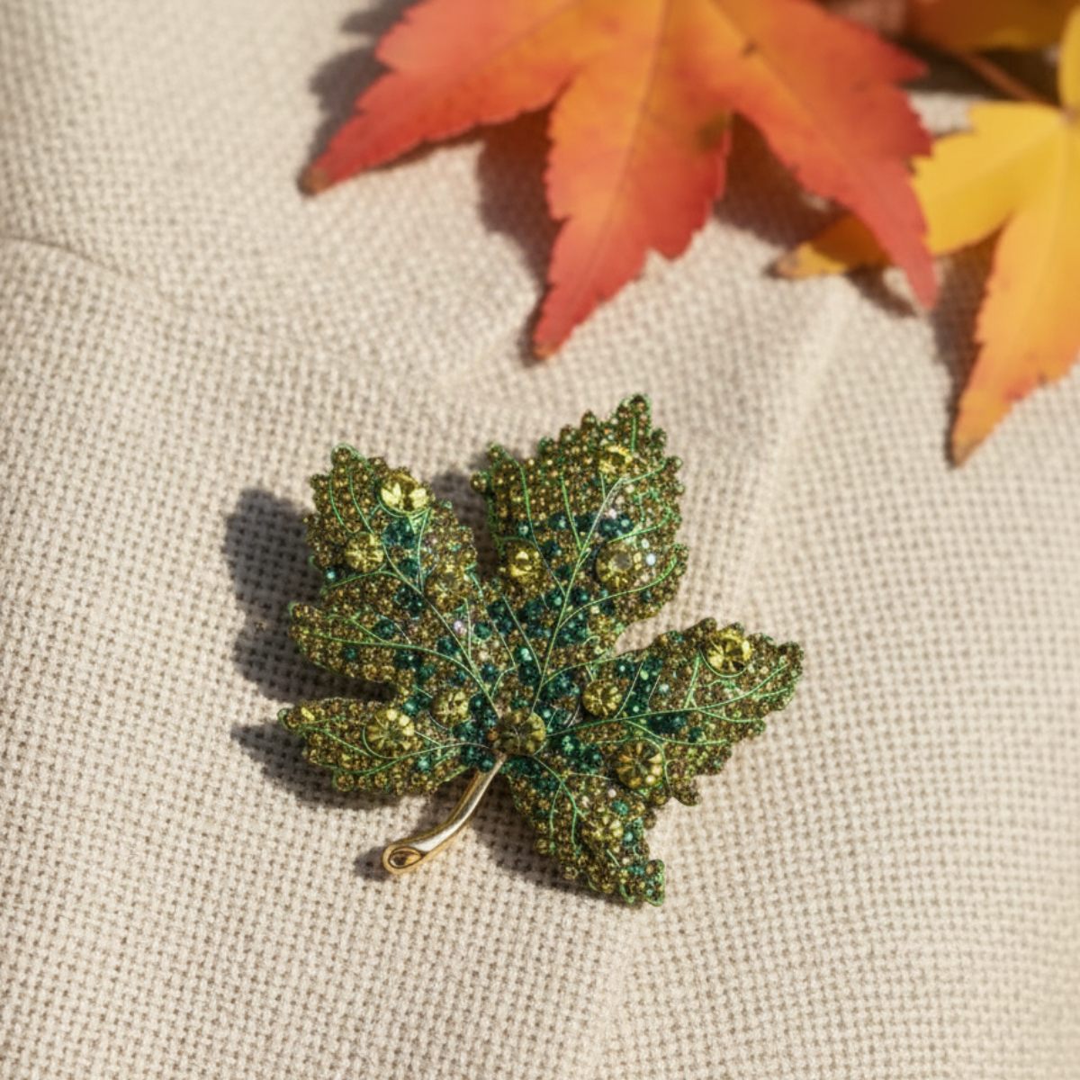 Green leaf-shaped brooch on a brown textured fabric background with autumn leaves. Green Maple Leaf Crystal Brooch Pin 2" | Fall Rhinestone Leaf Pin