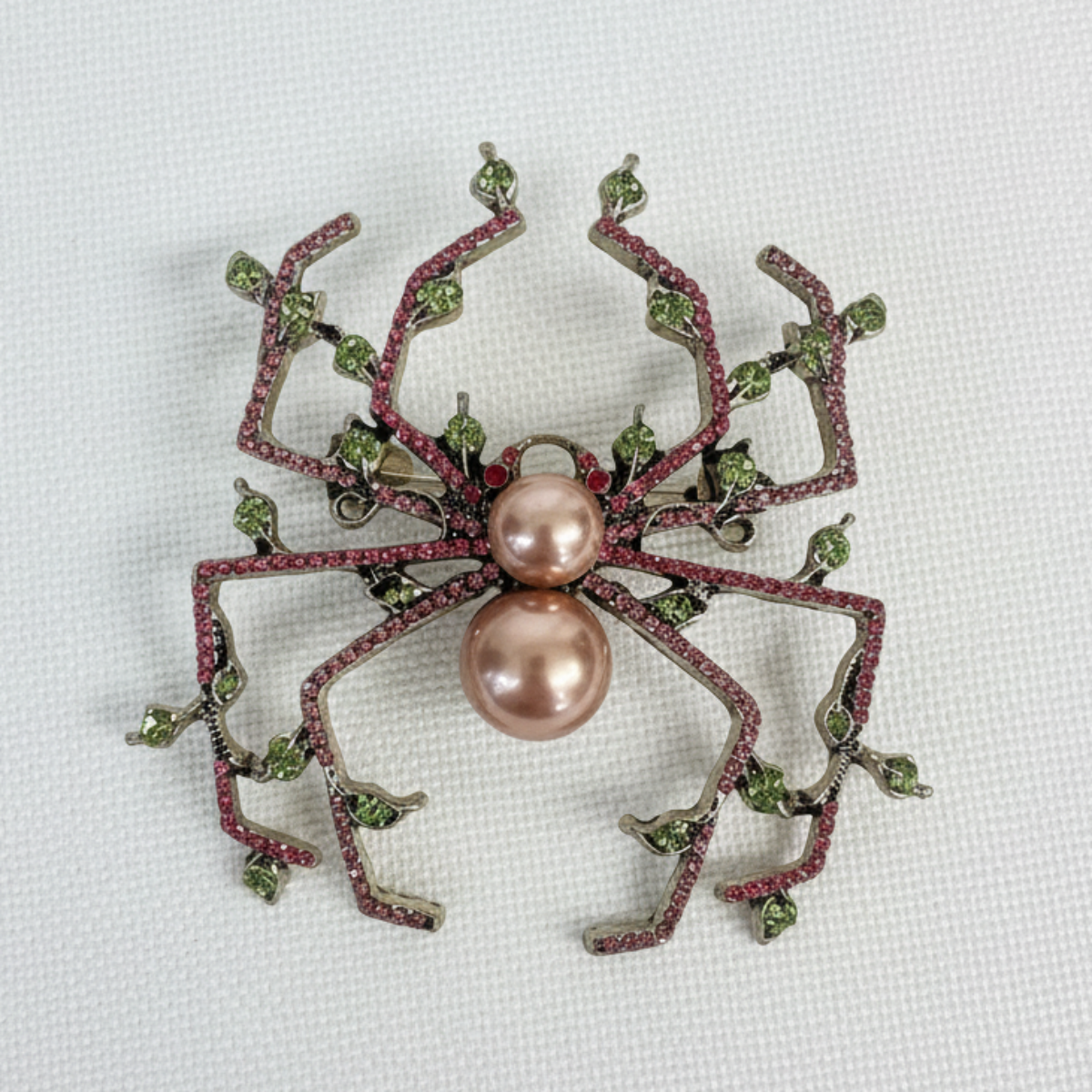 Spider-shaped brooch with pink pearl center and green gemstones on a white background,