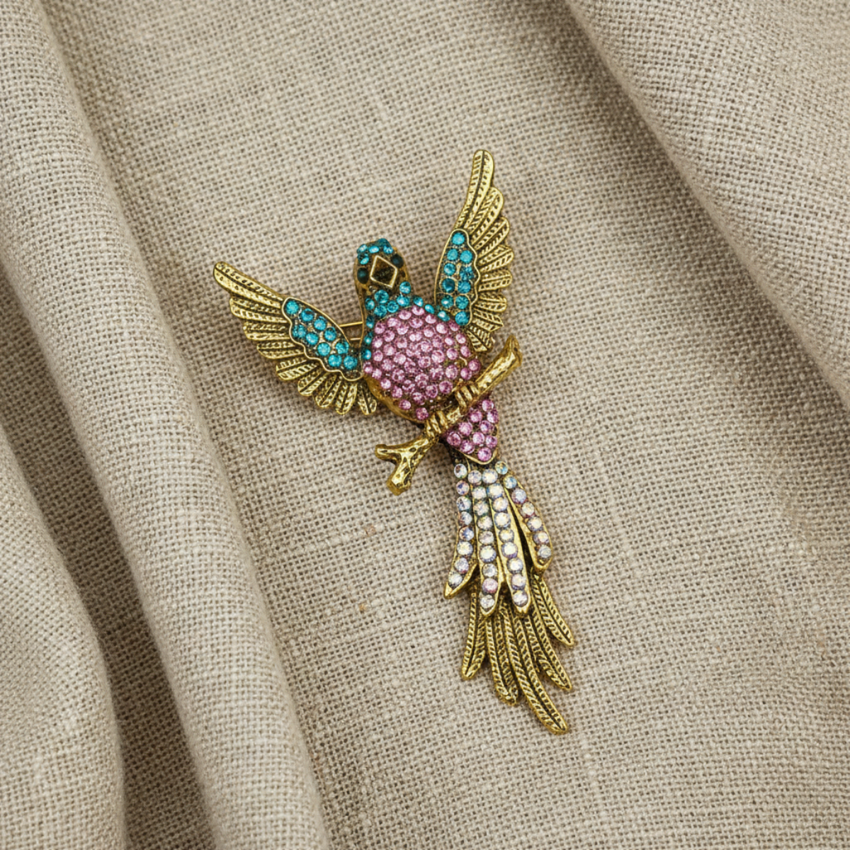 A gold bird brooch embellished with pink rhinestones, turquoise blue stones, and white pearls, resting on a textured beige linen fabric background.