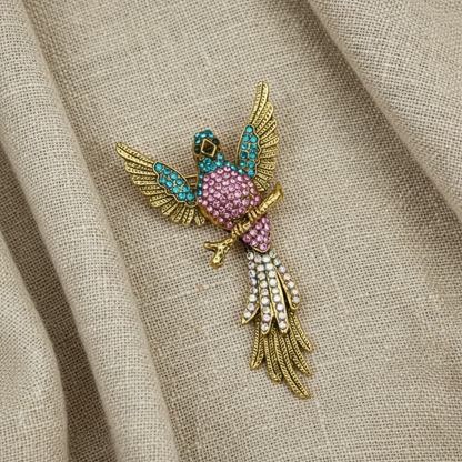 A gold bird brooch embellished with pink rhinestones, turquoise blue stones, and white pearls, resting on a textured beige linen fabric background.