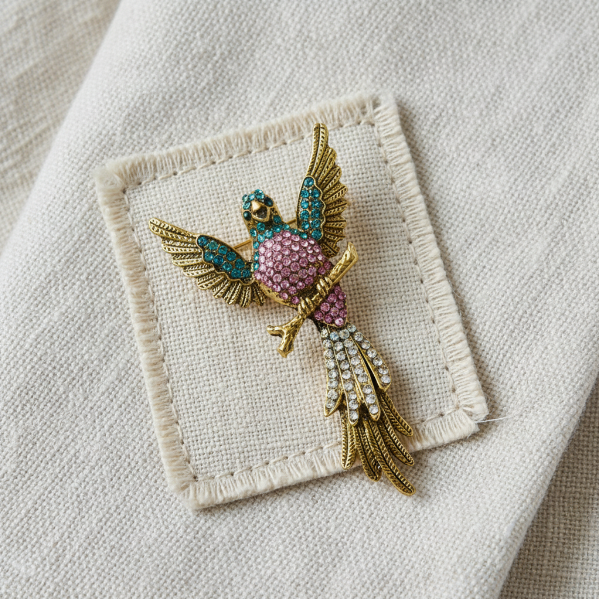 A gold bird brooch with turquoise and pink rhinestone accents, displayed on a beige embroidered fabric patch.
