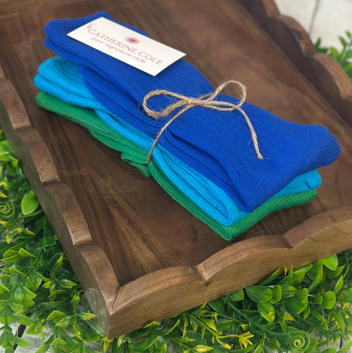 Stack of blue and green socks tied with twine on a wooden tray with a brand label, surrounded by green foliage.