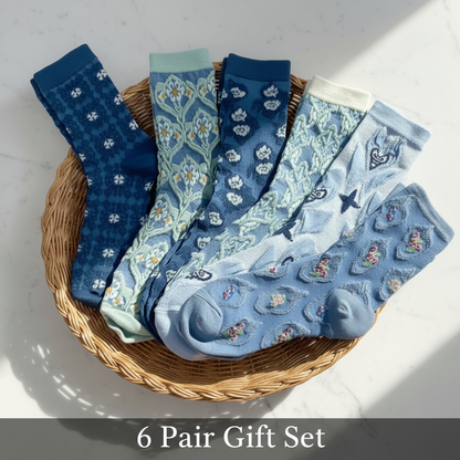 Six pairs of patterned baby socks arranged in a round woven basket on a light surface. The set includes various blue and navy patterns such as florals, stripes, and stars, with one pair in light blue and one pair in mint green.