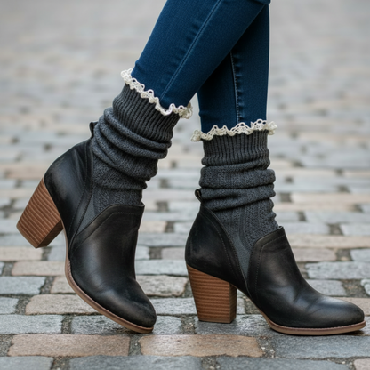 Black ankle boots with brown soles worn with blue jeans and gray leg warmers on a stone pavement.