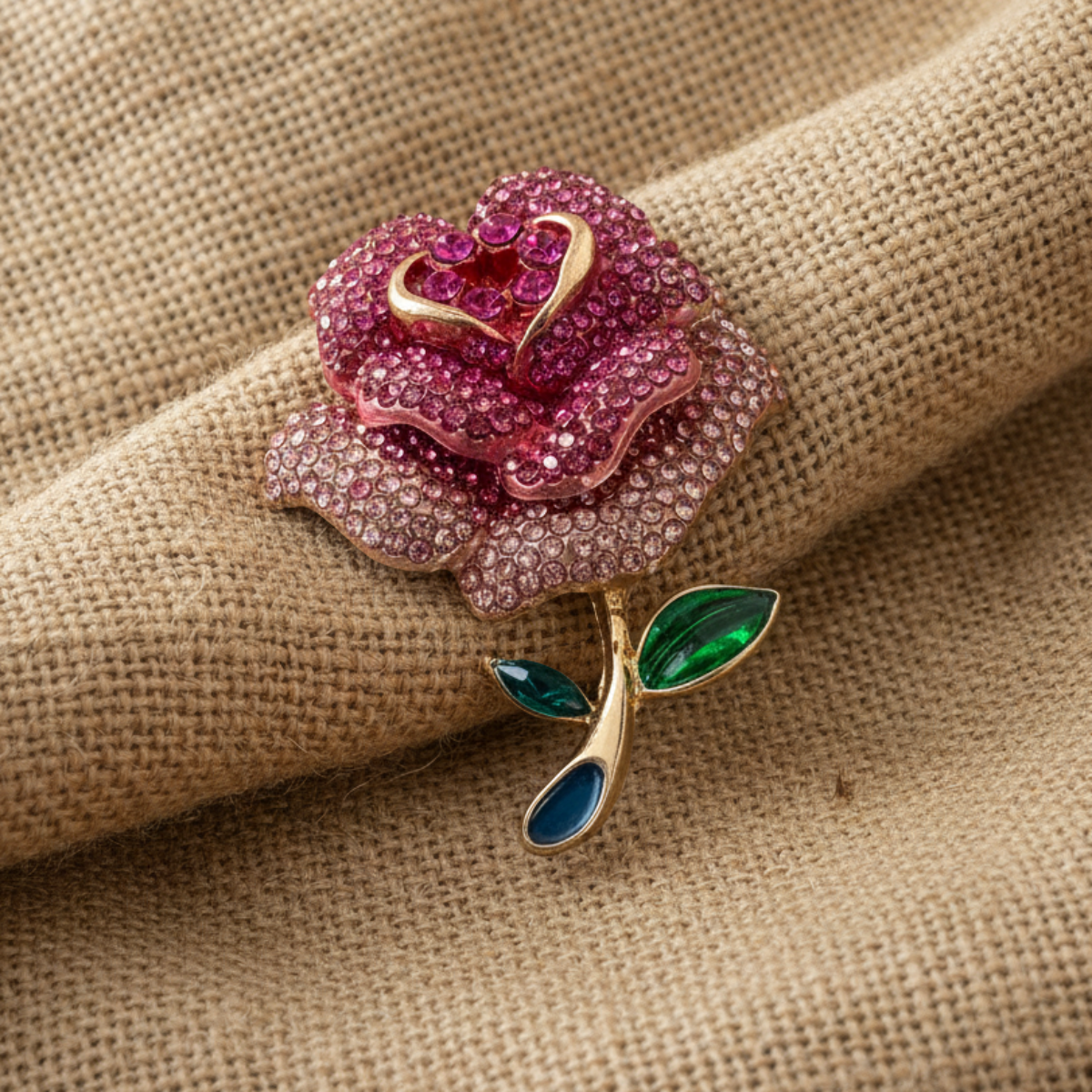 Pink rose-shaped brooch with gold and green accents on a beige fabric background