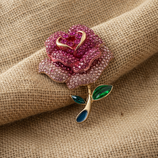 Pink rose-shaped brooch with gold and green accents on a beige fabric background
