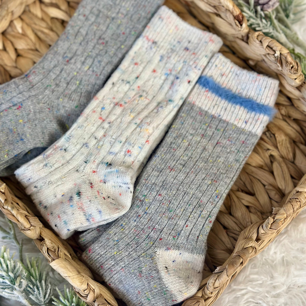 Three pairs of socks in gray and multicolor on a woven basket with plants in the background, Cozy grey mix patterned socks - 3 pair gift set + wish bracelet