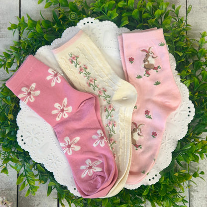 Three pairs of pink socks with floral and bunny designs on a white lace doily with green leaves in the background.