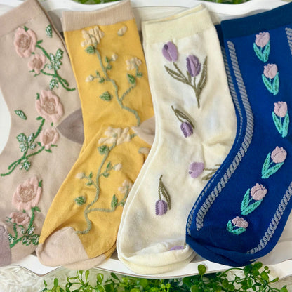 Four pairs of embroidered socks in different colors on a white surface with greenery.  Floral Flower Ankle Socks 4-Pack Bundle with Wish Bracelet - Women's 6-9