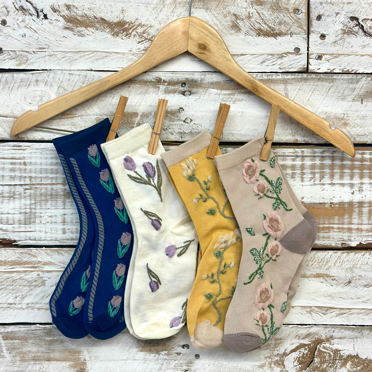 Four pairs of embroidered socks on a wooden hanger against a rustic wooden background. Floral Flower Ankle Socks 4-Pack Bundle with Wish Bracelet - Women's 6-9