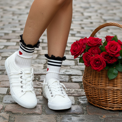 White sneakers with black and white striped socks featuring red hearts, next to a basket of red roses on a stone pavement.