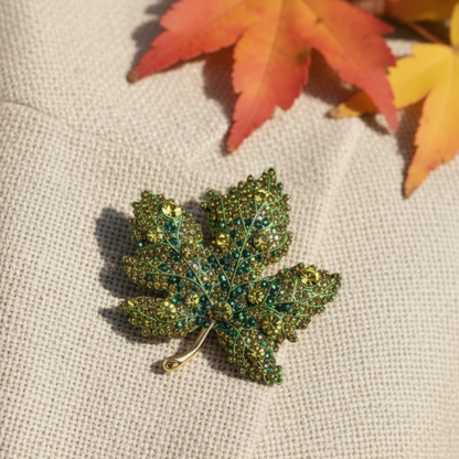 Green leaf-shaped brooch on a brown textured fabric background with autumn leaves. Green Maple Leaf Crystal Brooch Pin 2" | Fall Rhinestone Leaf Pin