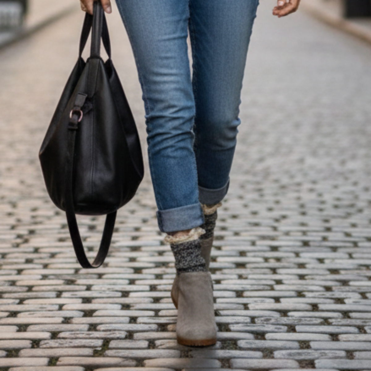 Person walking on a cobblestone street holding a black handbag, wearing blue jeans and brown shoes. salt n pepper organic socks