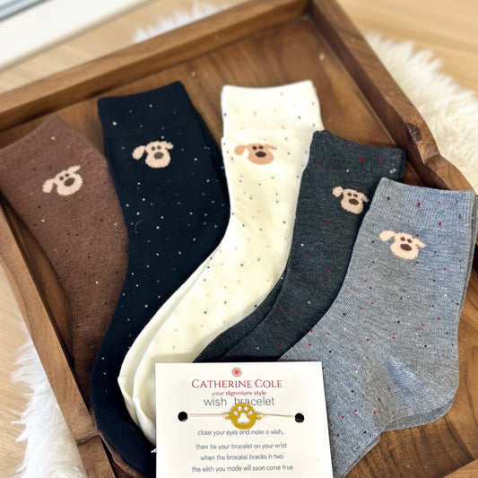 Four pairs of socks with dog patterns on a wooden surface, featuring a Catherine Cole bracelet card.