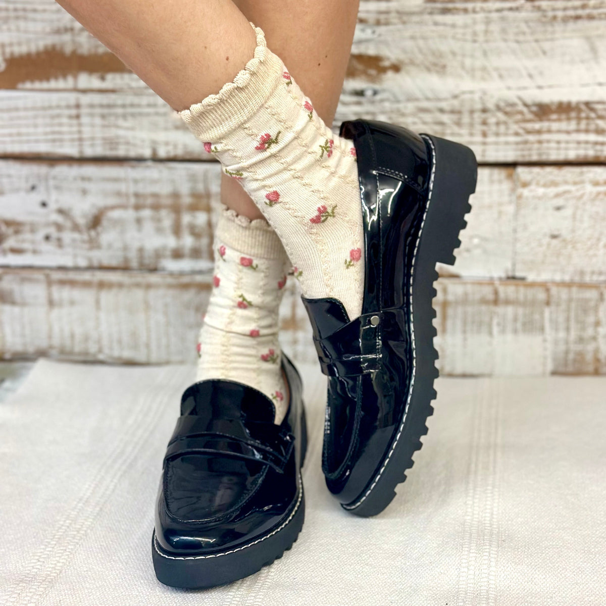 Black patent leather shoes worn with white socks featuring floral patterns on a textured surface.