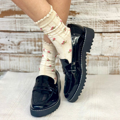 Black patent leather shoes worn with white socks featuring floral patterns on a textured surface.