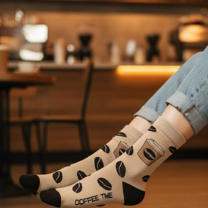 Person wearing socks with coffee-themed design in a blurred cafe setting.  The Coffee Lover Gift Set (2-Pack Socks + Coffee Cup Earrings)