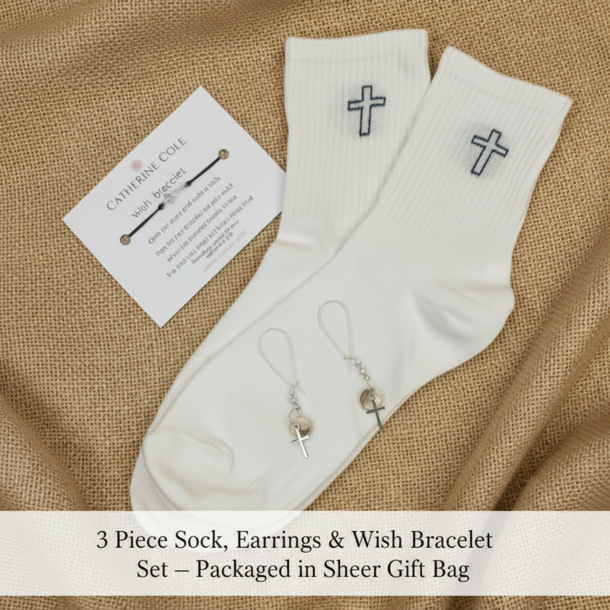 A pair of white ribbed crew socks with small black cross embroidery on the cuffs, displayed on a light fabric background alongside a small branded card and a pair of dangling earrings.