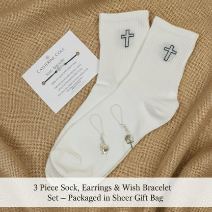 A pair of white ribbed crew socks with small black cross embroidery on the cuffs, displayed on a light fabric background alongside a small branded card and a pair of dangling earrings.
