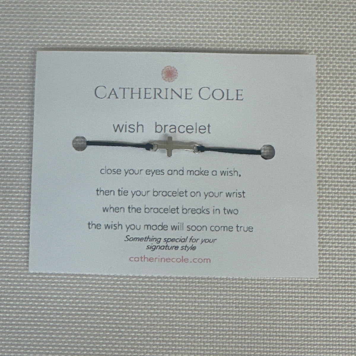 A Catherine Cole wish bracelet with a silver cross charm and black cord, displayed on its white branded card against a textured light gray background.