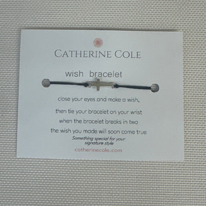 A Catherine Cole wish bracelet with a silver cross charm and black cord, displayed on its white branded card against a textured light gray background.