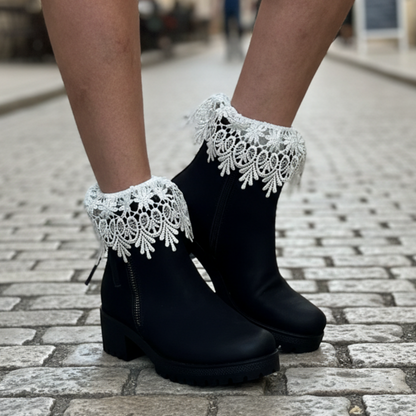 Black ankle boots with white lace trim at the top, shown on a person's feet standing on a cobblestone street. cute lace socks for short ankle boots by Catherine cole
