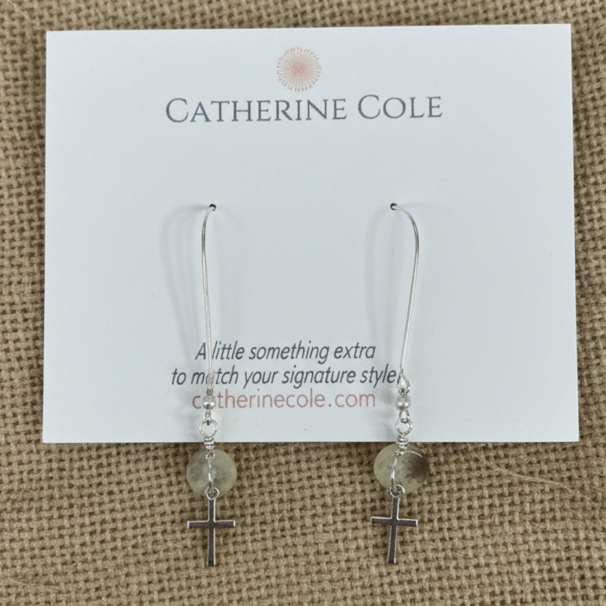 A pair of silver-toned dangle earrings with small pale yellow beads, displayed on a white Catherine Cole brand card against a light gray textured fabric background.