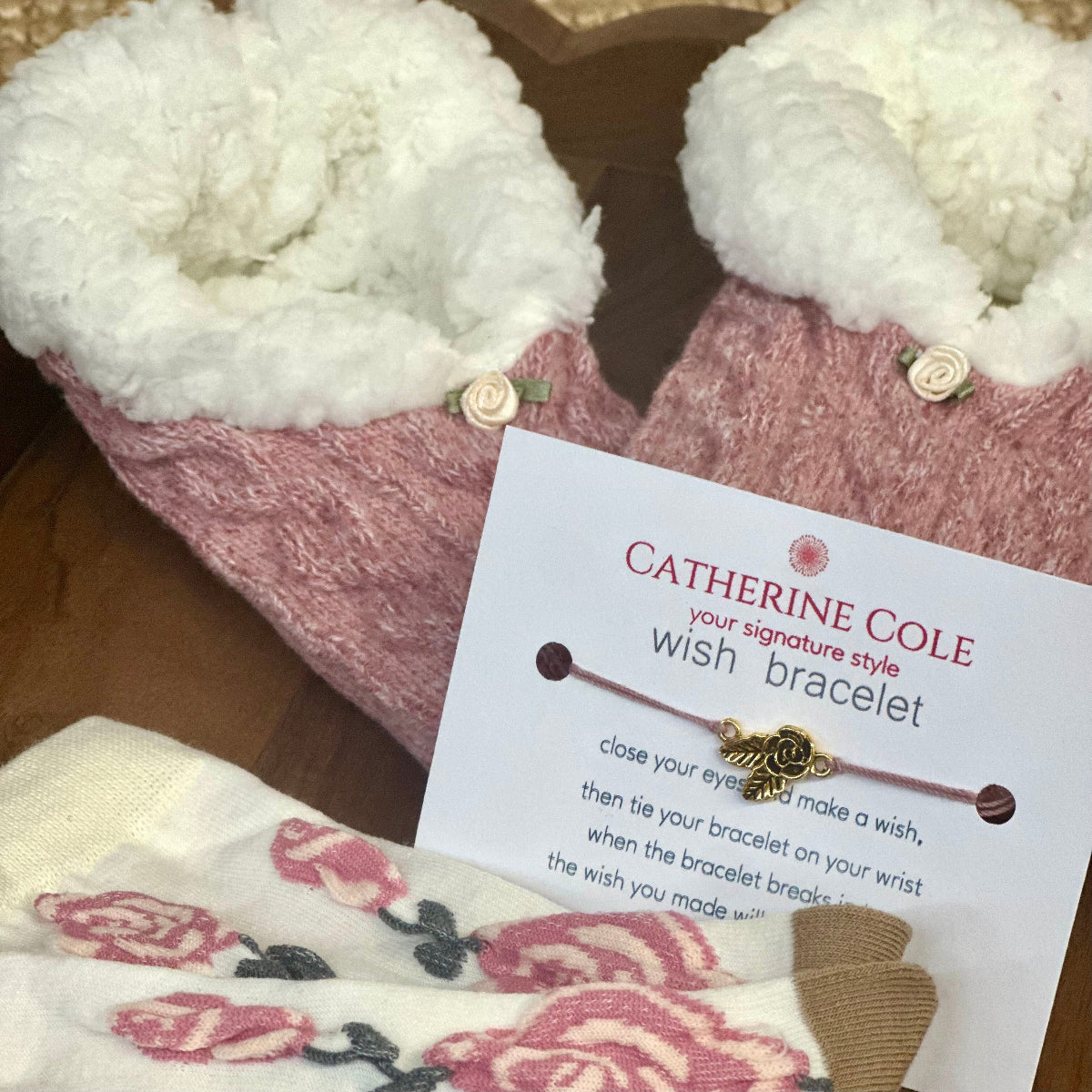 Pink slippers with white fur lining and a Catherine Cole wish bracelet card on a wooden surface.