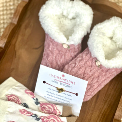 Pink slippers with white fur lining and a Catherine Cole bracelet on a wooden surface.