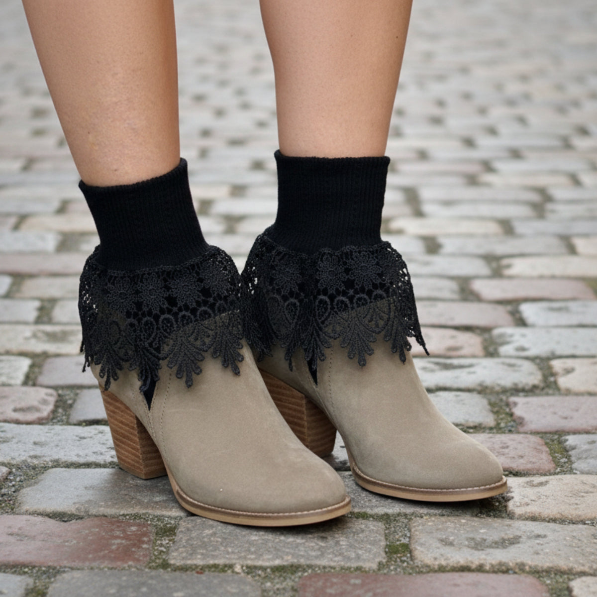 Beige ankle boots with black lace details worn on a cobblestone street with autumn leaves.