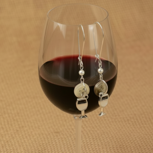 Silver earrings with pearls hanging over a glass of red wine on a brown textured background
